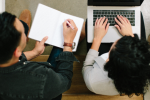 two people. One is typing on a computer. One is holding a paper.